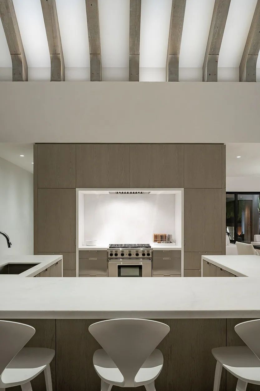 Kitchen with central island, bar stools, wood cabinetry, built-in oven, and exposed beams with natural light.
