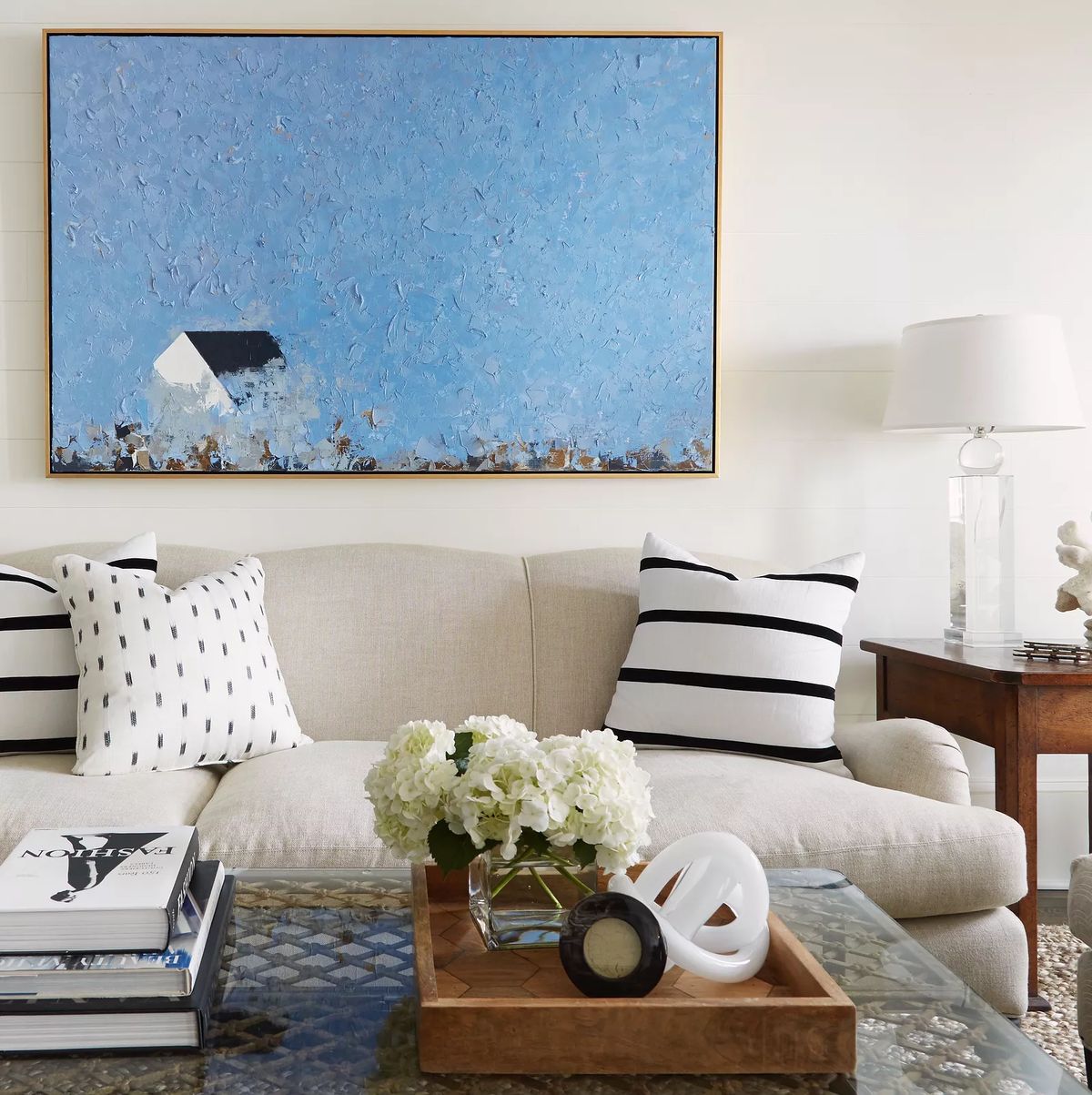 Living room with beige sofa, striped cushions, wooden table, glass coffee table, and large blue artwork