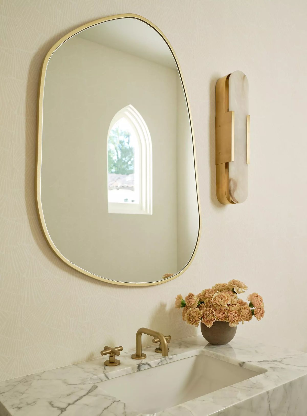 Powder room with marble countertop, gold faucet, oval mirror, dried flowers, wall sconce, and arched window.