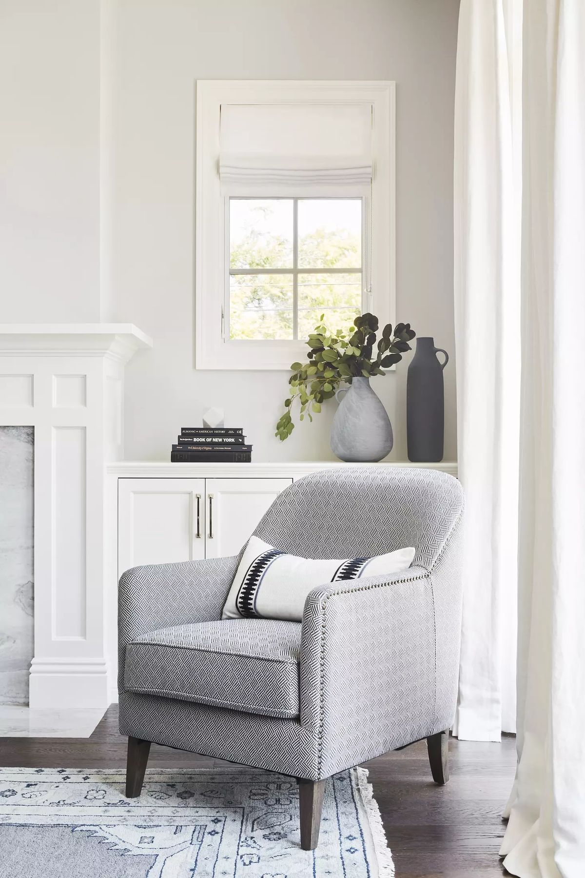 Living room with gray armchair, white cabinet, books, vase, and window with treatments for natural light.