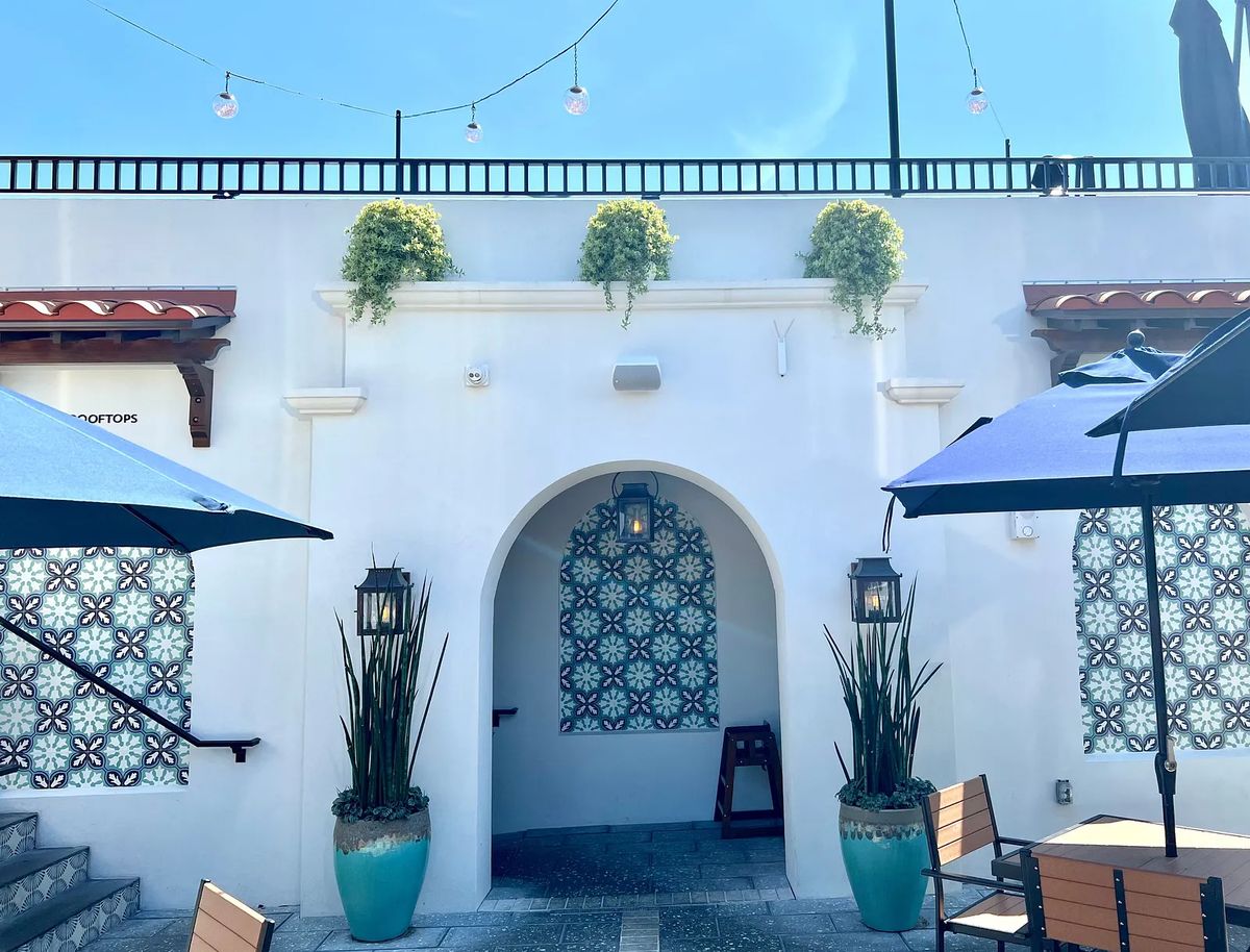 Outdoor deck with blue umbrellas, wooden tables, chairs, white walls, and decorative tiles.