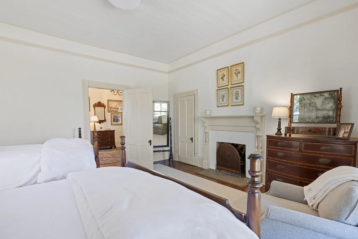 Bedroom with two white beds, wooden dresser, armchair, fireplace, and a door leading to another room.