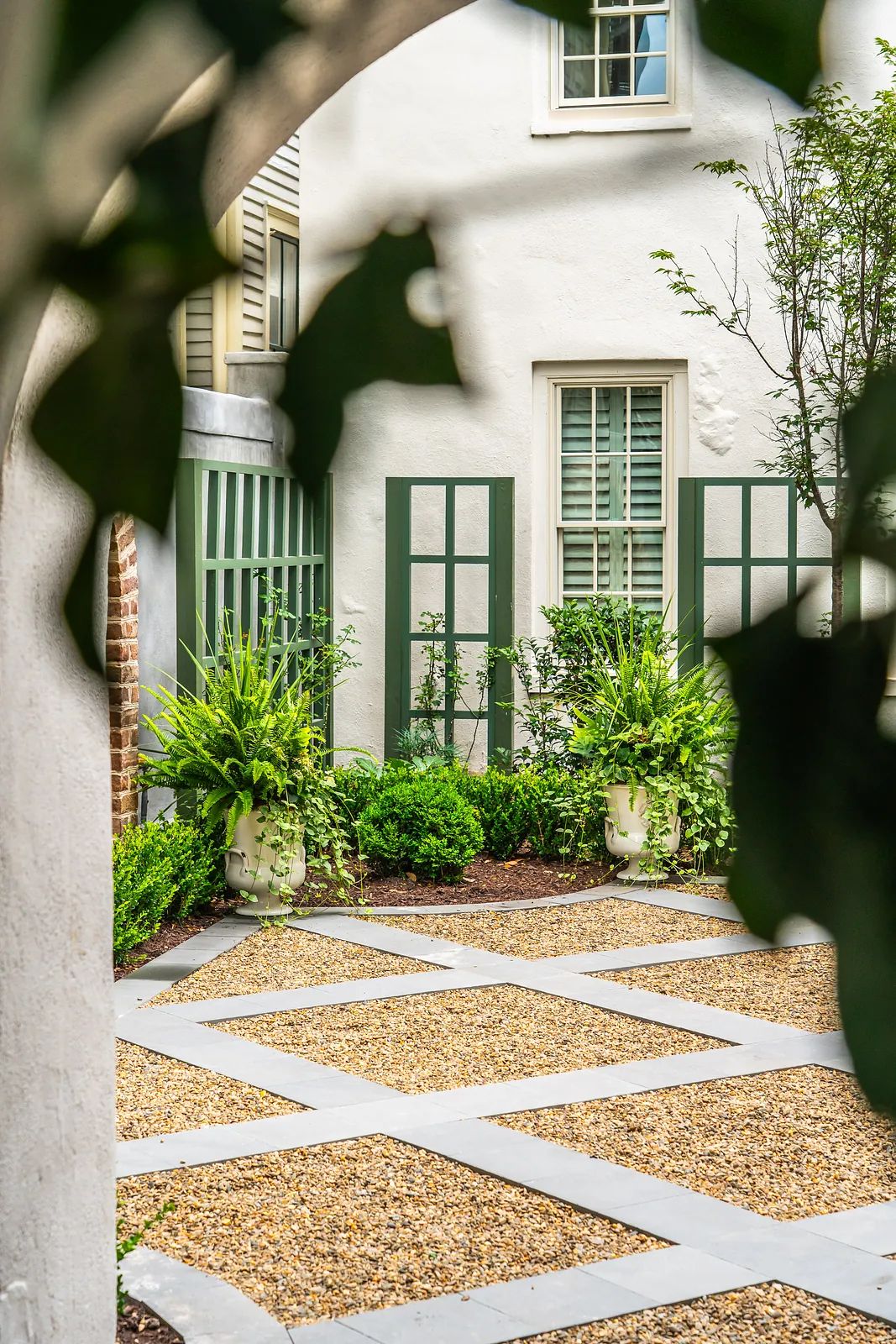 Garden with gravel path, potted plants, shrubs, and green shutters on windows.
