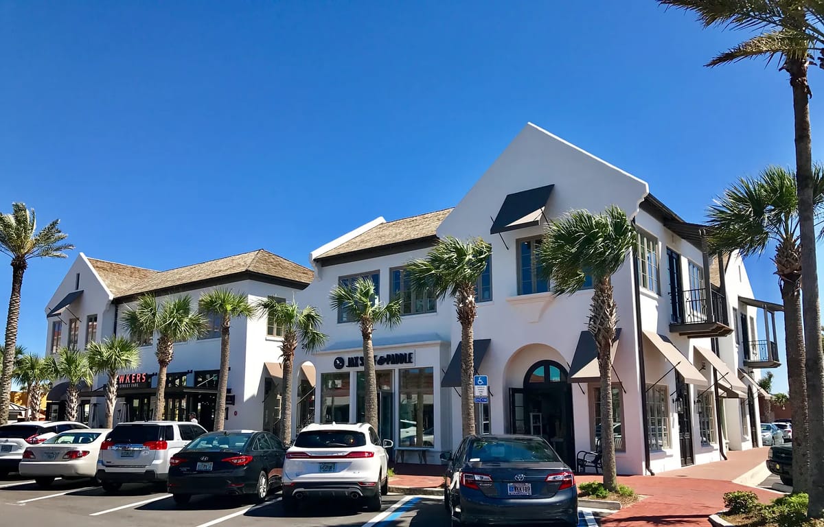 Exterior front view of a commercial building complex with white walls, tan roofs, palm trees, and parked cars.