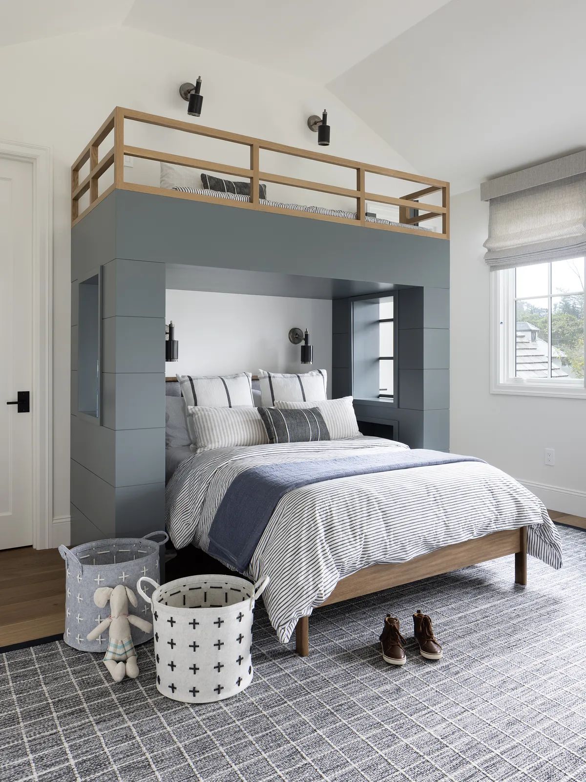 Bedroom with lofted bed, gray duvet, striped pillows, baskets, and patterned area rug with natural light