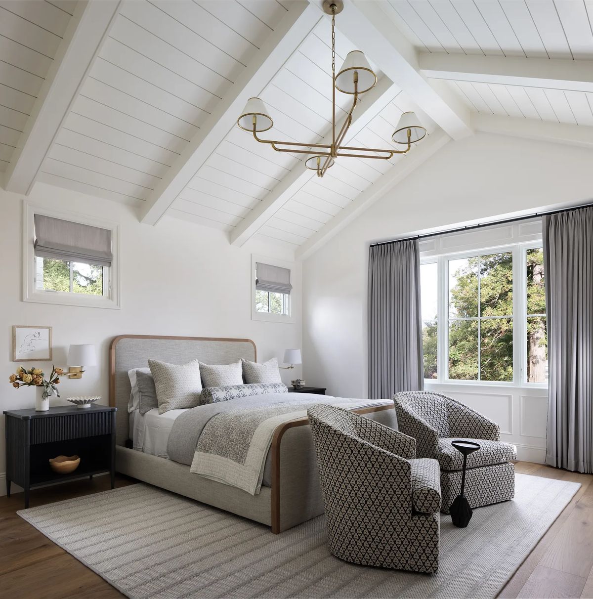 Bedroom with gray upholstered bed, patterned armchairs, large windows, and vaulted ceiling.