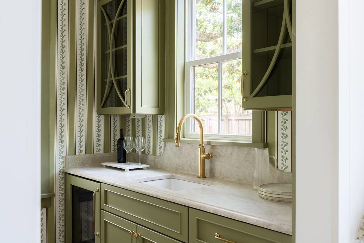 Kitchen area with green cabinetry, marble countertop, gold faucet, wine glasses, and patterned wallpaper.