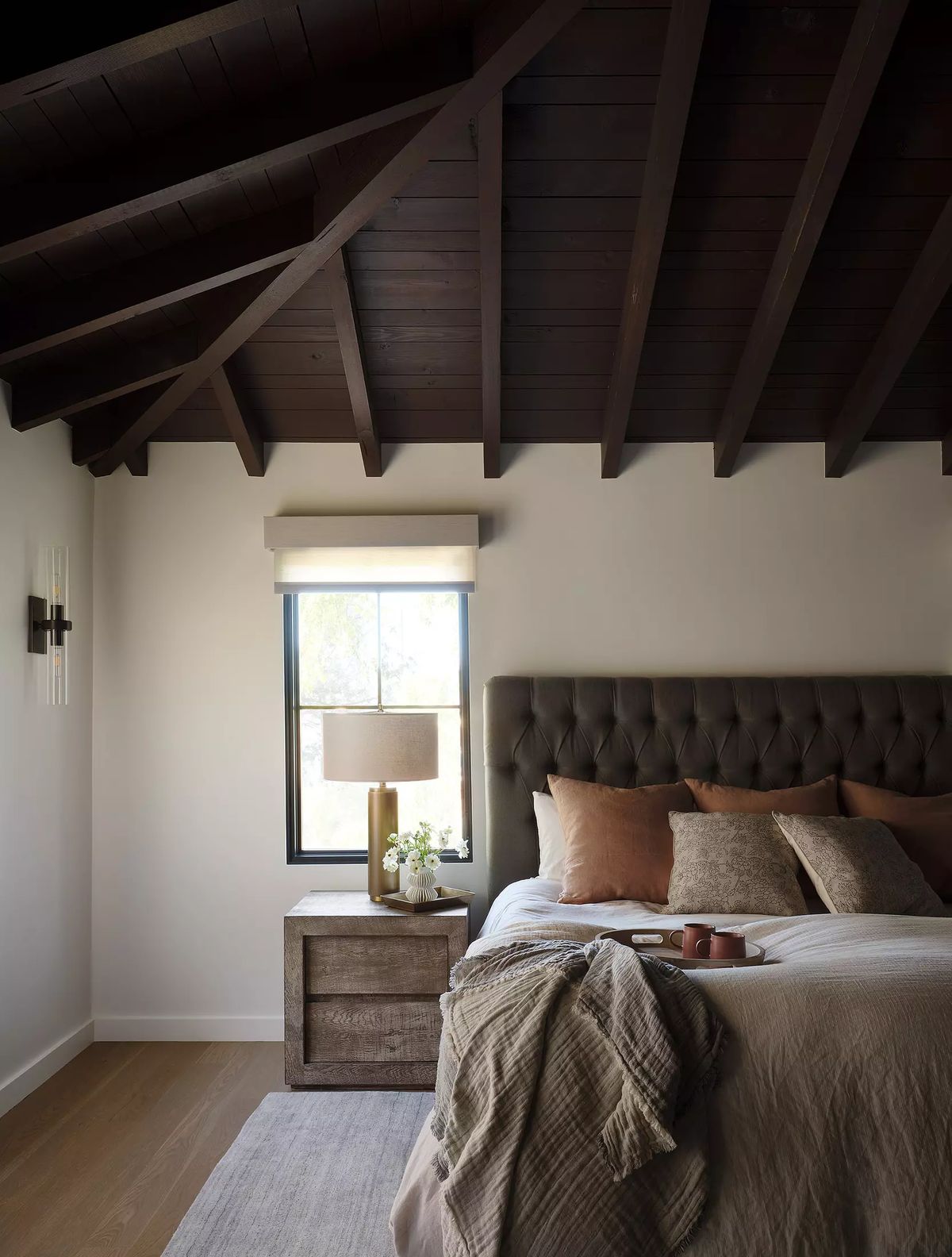 Bedroom with tufted headboard bed, nightstand, table lamp, vase, and exposed wooden beams.