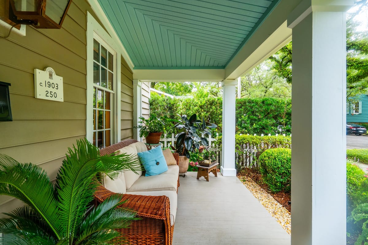 Porch with beige sofa, blue cushions, wicker chair, side table, and surrounding greenery.