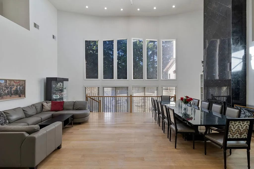 Dining room with long glass table, black chairs, sectional sofa, large windows, and decorative elements.