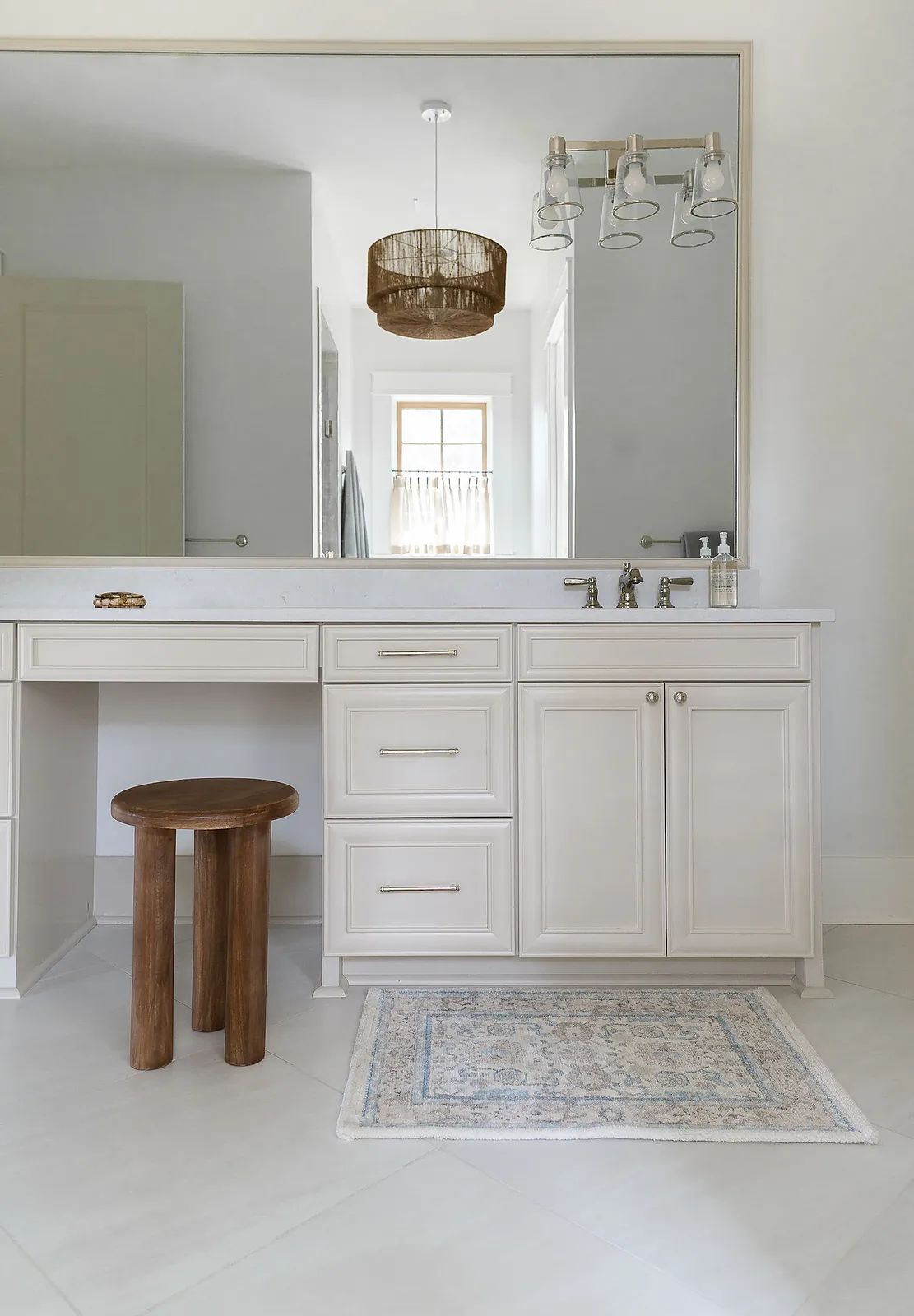 Bathroom with beige cabinetry, white countertop, wooden stool, patterned rug, large mirror, and white walls.