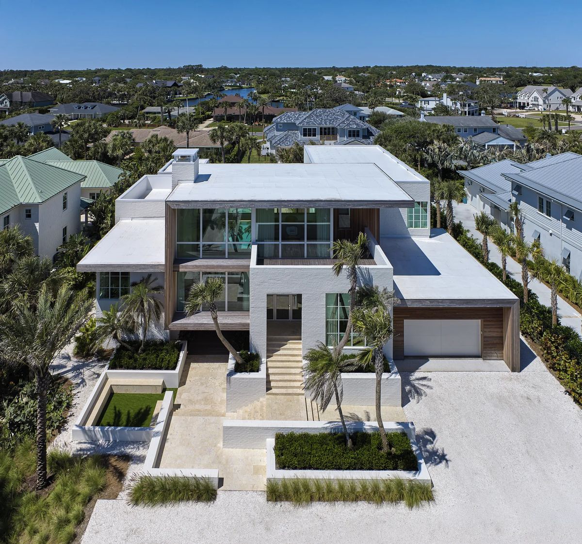 Modern house exterior with large windows, flat roof, palm trees, and landscaped area with a water feature.