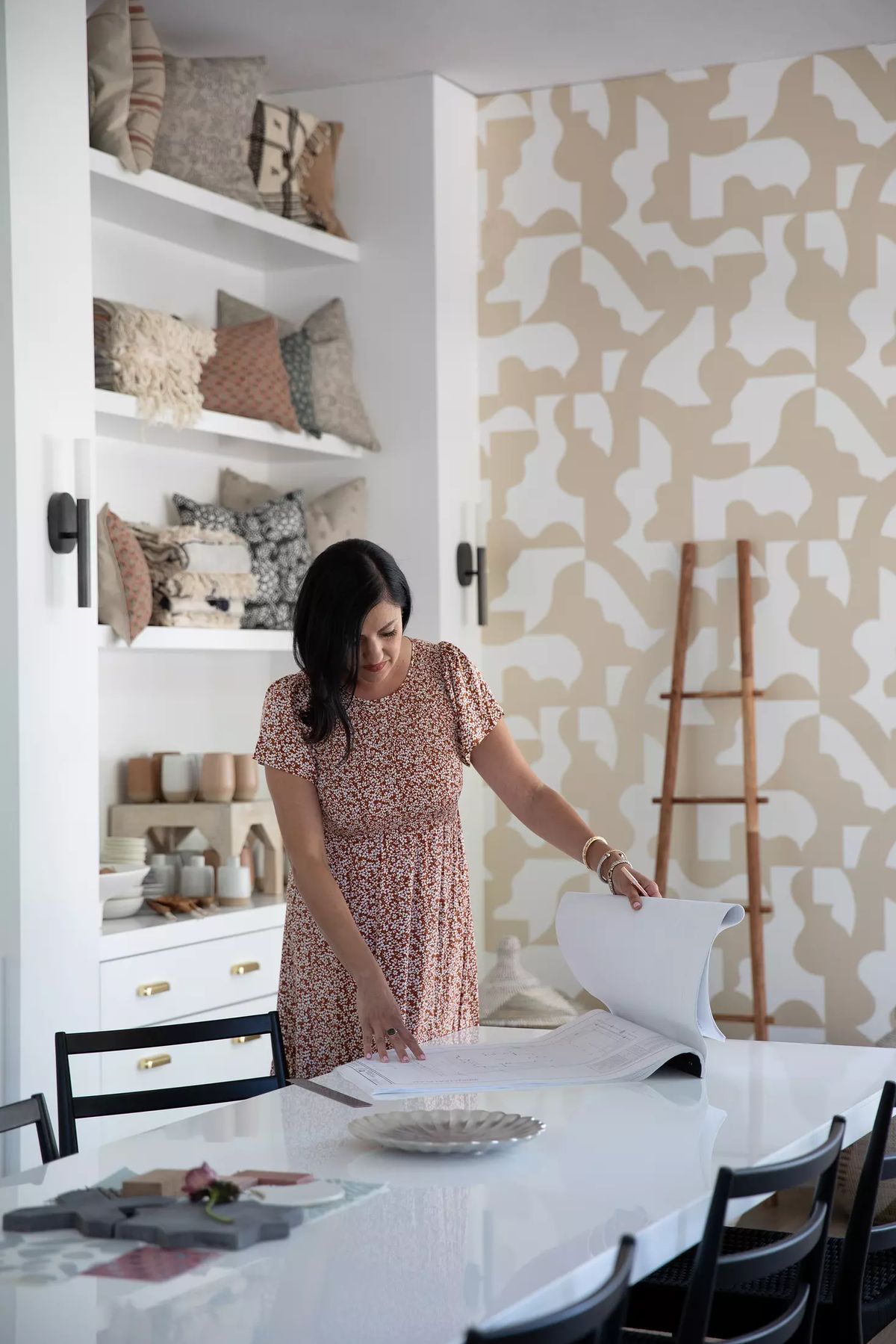 Dining room with white table, black chairs, shelves of decor, patterned wallpaper, and wooden ladder.