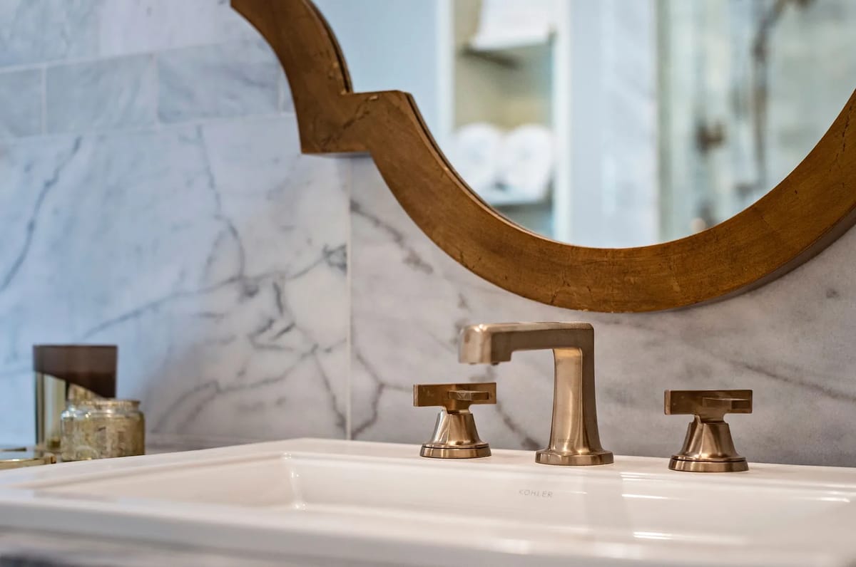 Bathroom with white sink, gold fixtures, round mirror, and marble backsplash.