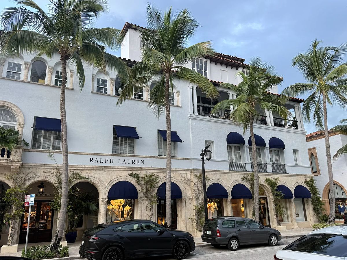 Building facade with light blue exterior, arched windows, blue awnings, and palm trees in front.
