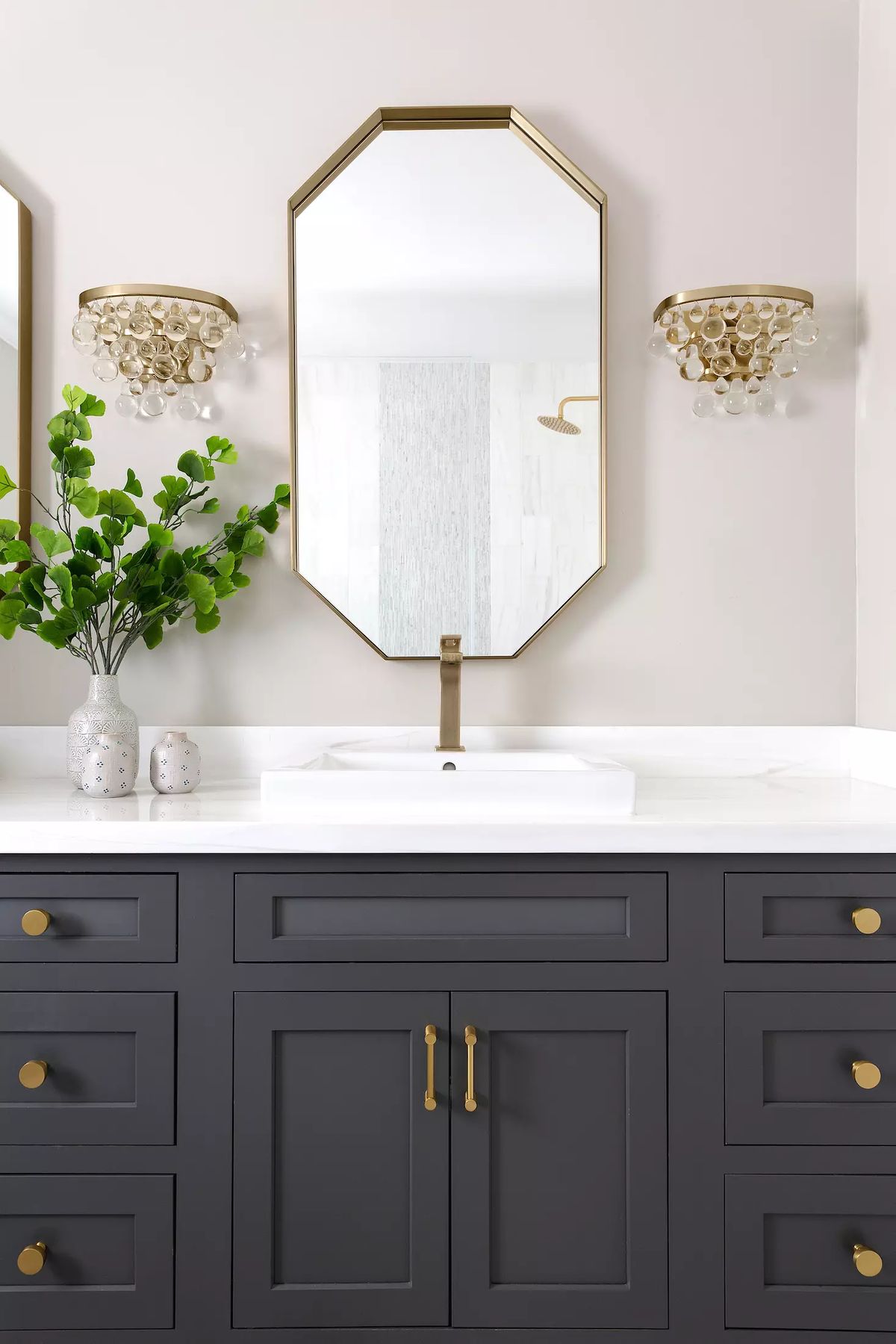 Bathroom with dark gray vanity, white sink, octagonal mirror, wall sconces, and decorative vase with foliage