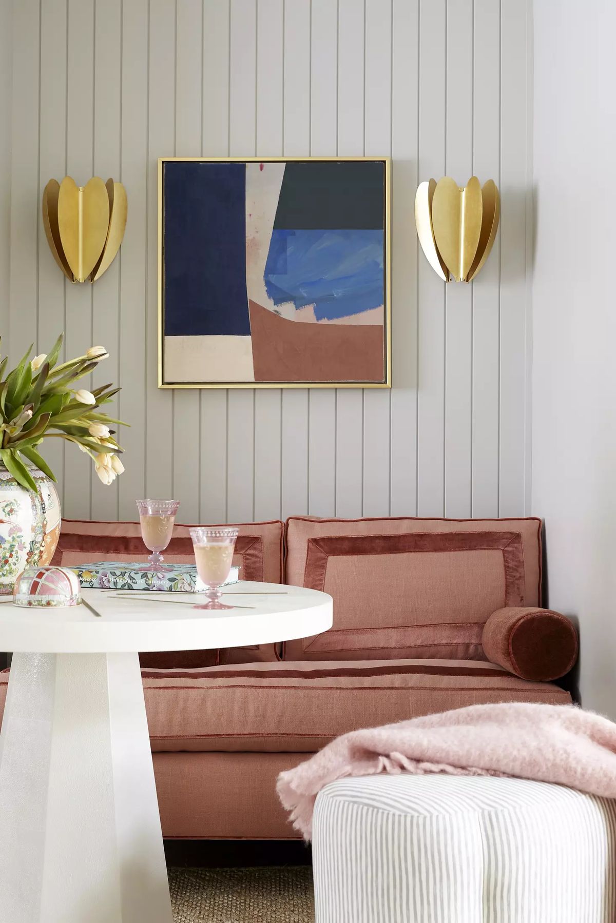 Living room with pink sofa, round white table, floral vase, abstract artwork, and gold wall sconces