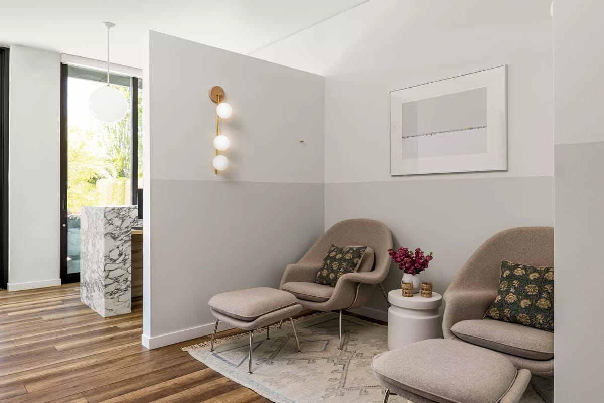 Seating area with grey armchairs, round side table, floral arrangement, and marble counter separating from kitchen.