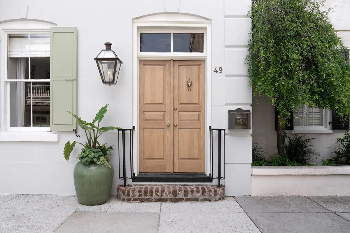Exterior front with wooden double doors, green shutters, lantern, planter, and brick walkway.