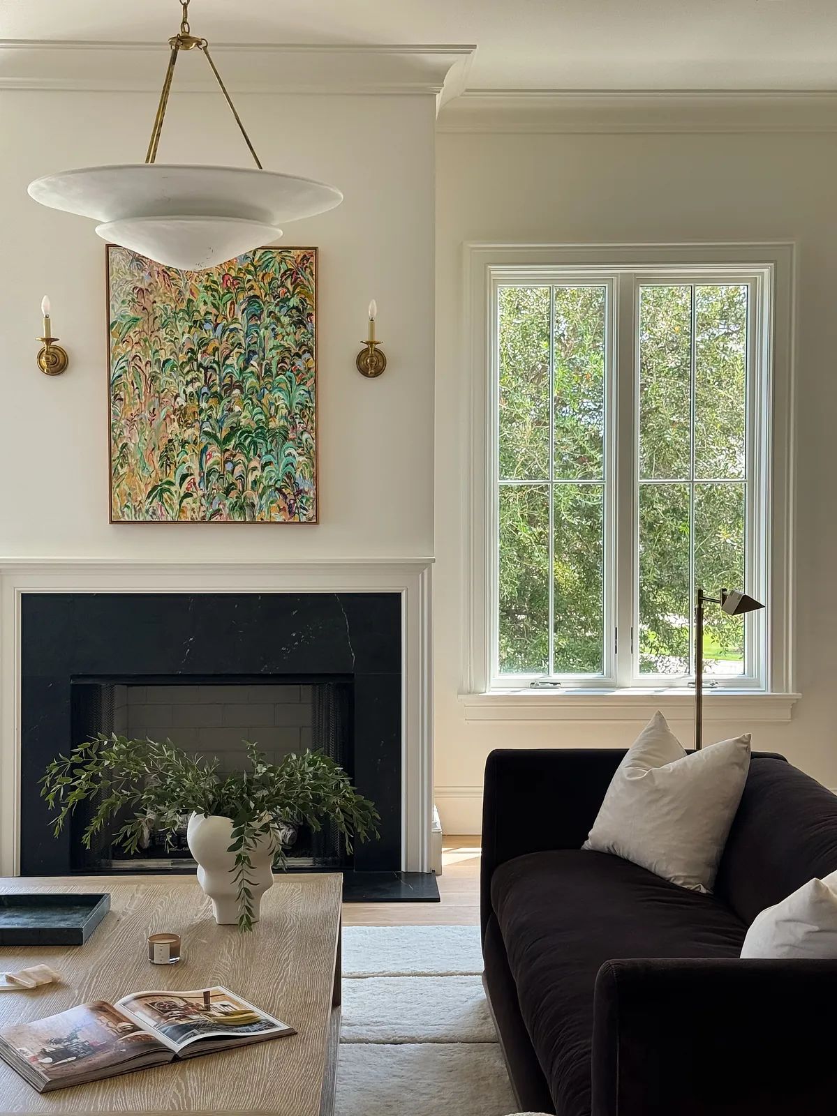 Living room with dark sofa, coffee table, side table, artwork, marble fireplace, and large windows with greenery view.