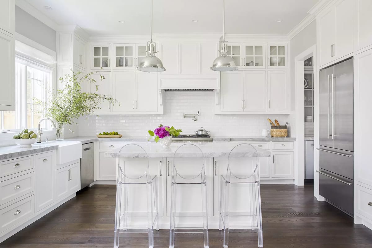 Kitchen with white cabinetry, marble island, clear stools, stainless steel appliances, and large windows for natural light.