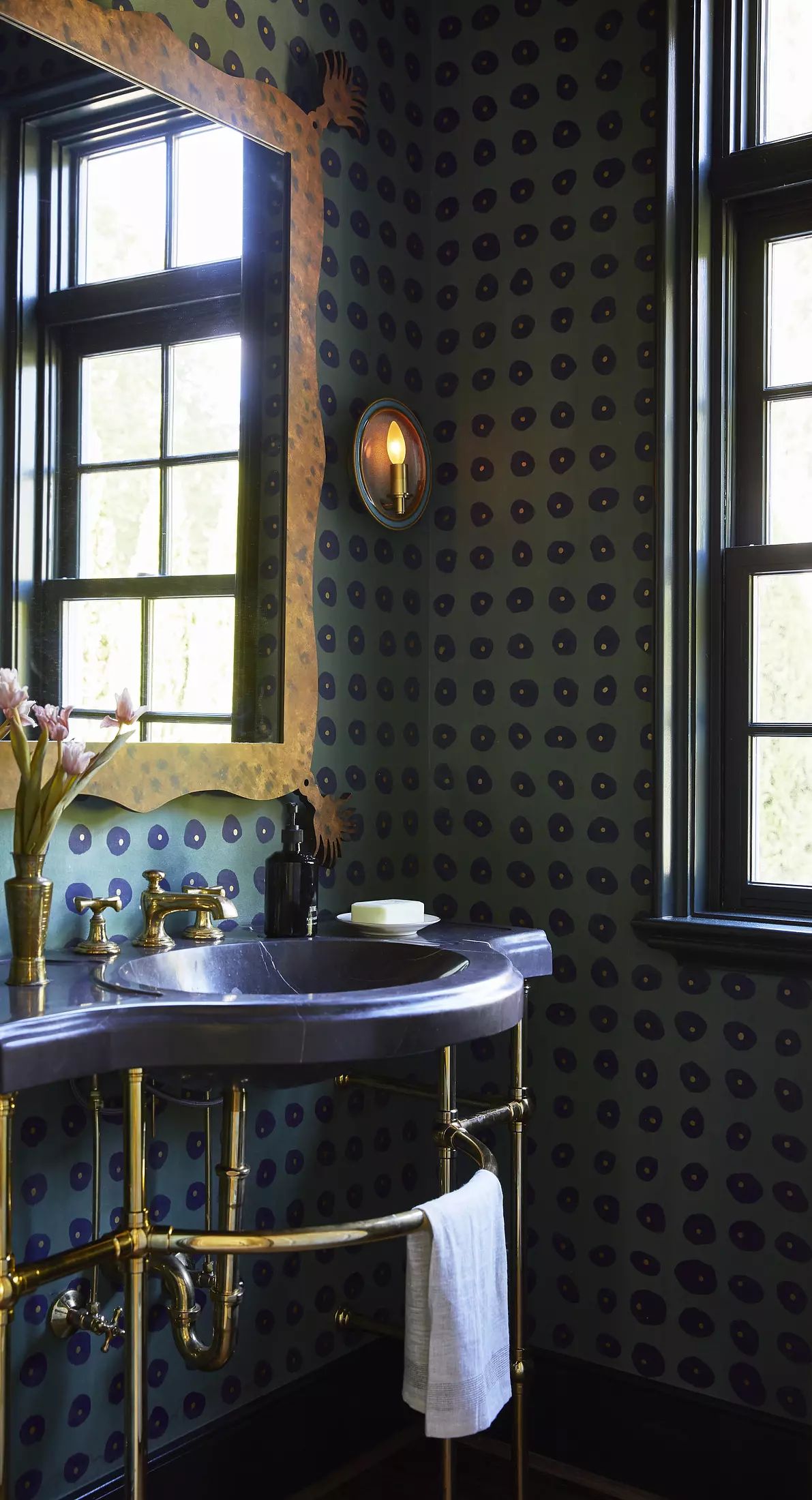 Bathroom with round sink, brass fixtures, patterned wallpaper, two windows, and a flower vase.