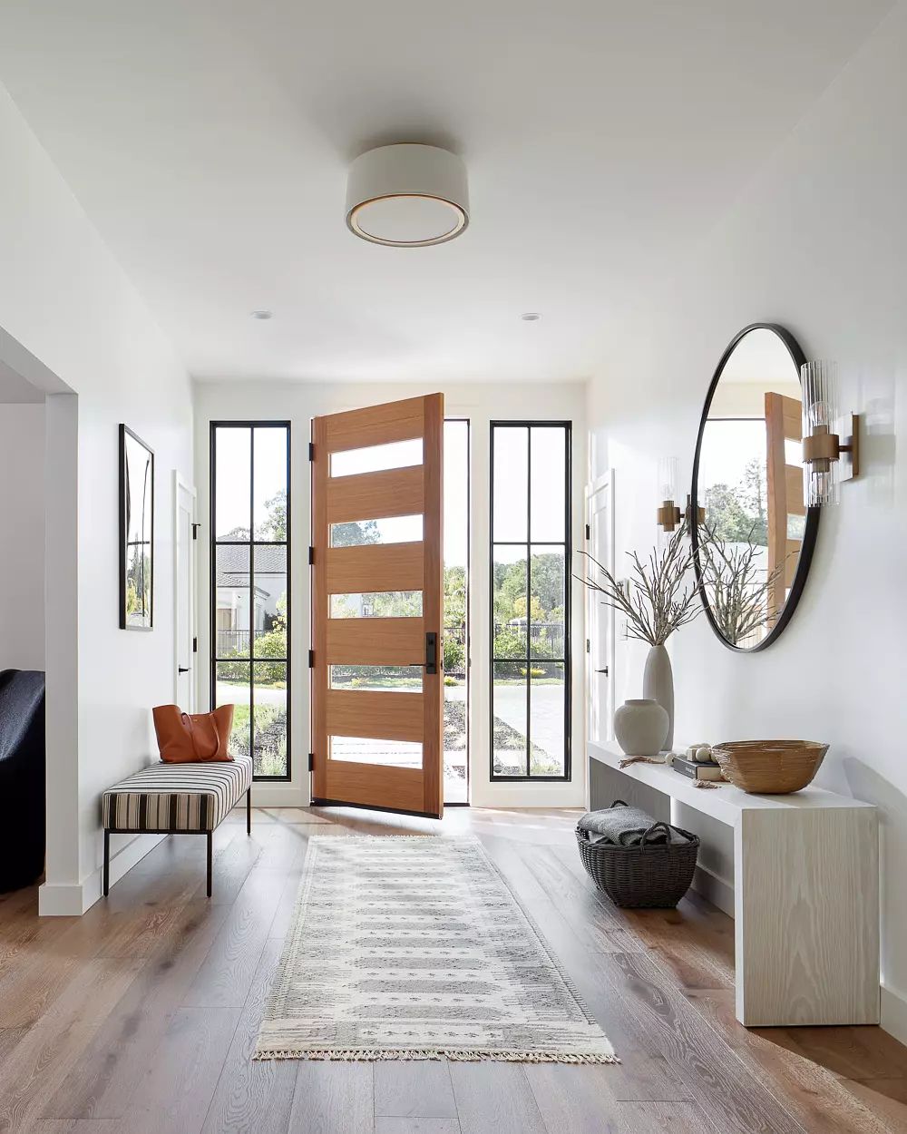 Foyer with wooden front door, windows, round mirror, console table, striped bench, and patterned rug.