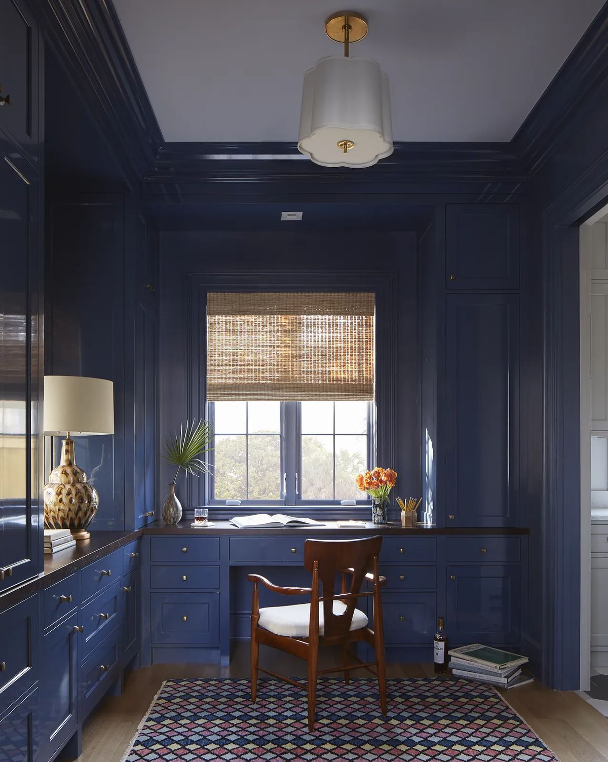 Office with blue paneled walls, built-in desk, wooden chair, colorful rug, and a vase with flowers