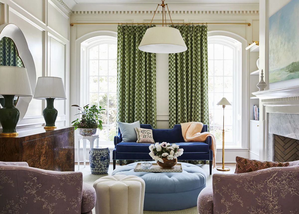 Living room with blue sofa, round ottoman, purple armchairs, wood sideboard, and large windows with green curtains.
