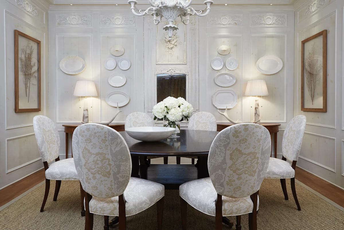 Dining room with round wooden table, six patterned chairs, decorative bowl, wall art, and lamps beside a mirror.