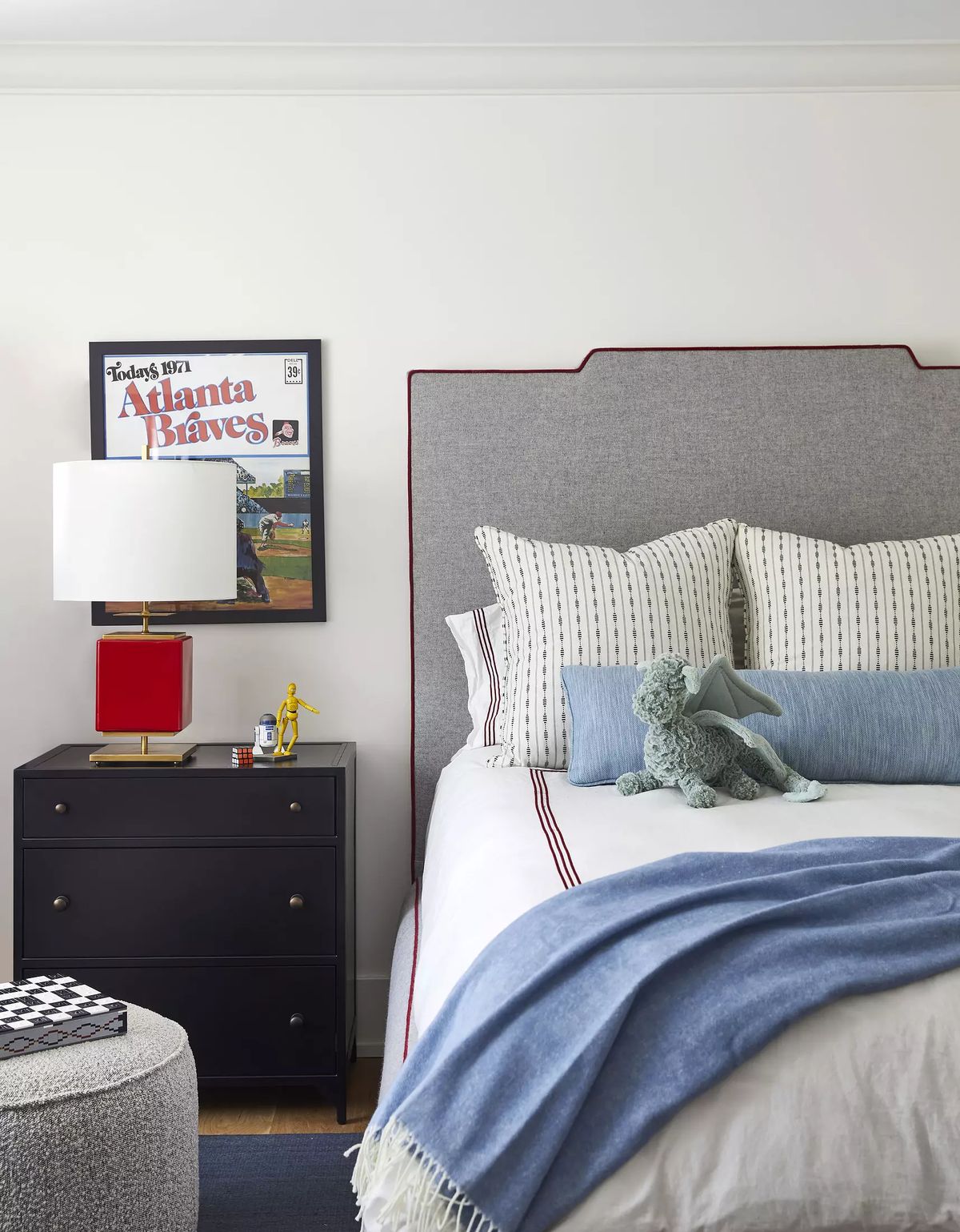 Bedroom with grey headboard, white bedding, red lamp, dark nightstand, blue throw, and framed poster.