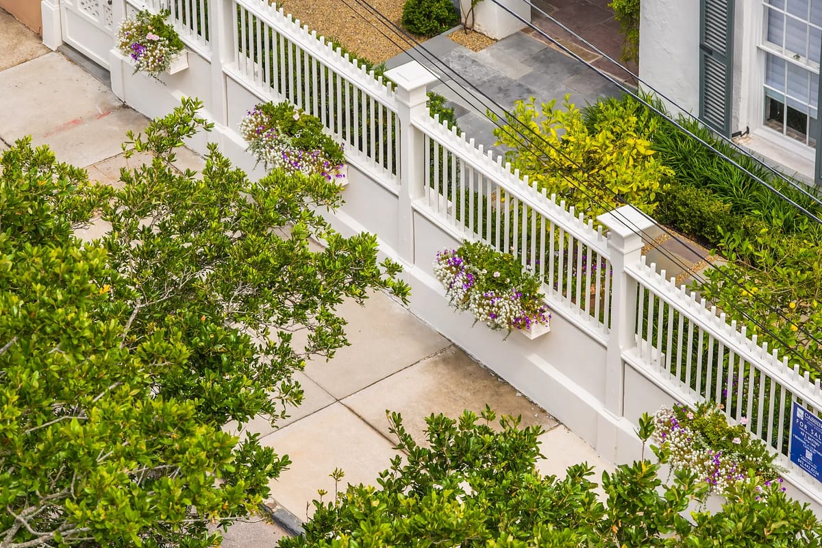 Exterior front view with white picket fence, hanging flowers, concrete sidewalk, and surrounding greenery.