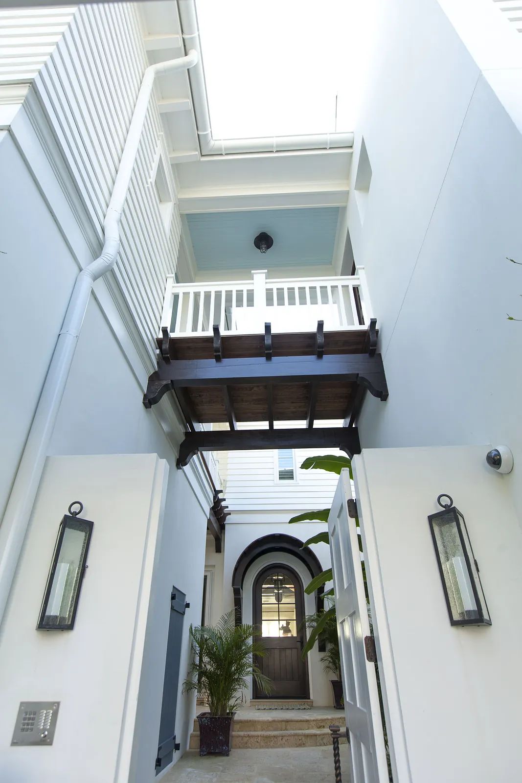 Exterior entrance with wooden balcony, arched doorway, two lanterns, and a palm plant.