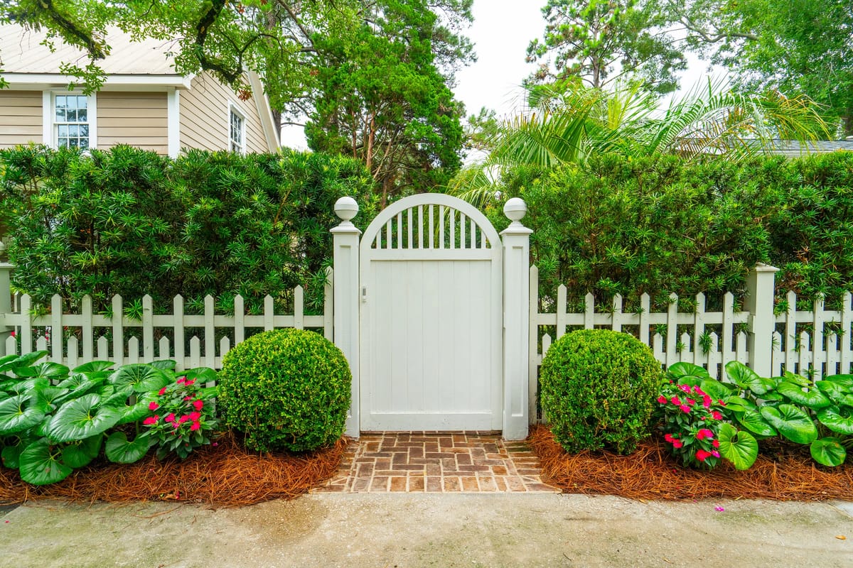 Exterior front yard with white picket fence, gated entry, circular shrubs, and pink flowering plants.