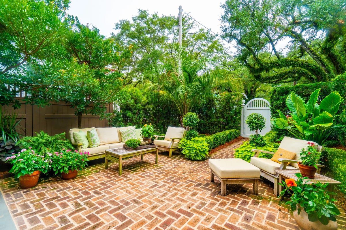 Garden with brick pavers, sofas, coffee table, and various plants including ferns and flowering plants.