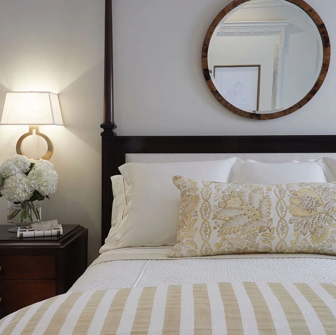 Bedroom with four-poster bed, nightstand with lamp and flowers, round mirror, and striped bedding