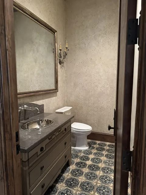Powder room with gray vanity, sink, toilet, mirror, decorative sconces, and patterned tile floor.
