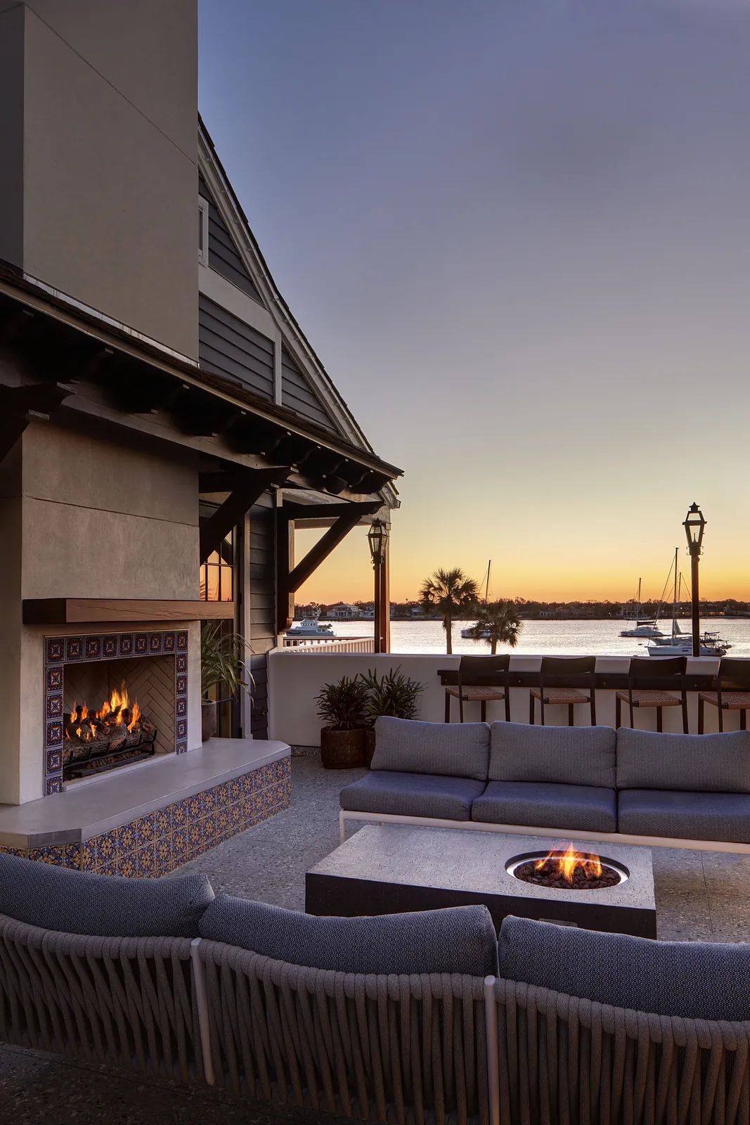 Deck with sectional sofa, fire pit table, tiled fireplace, and views of watercraft at sunset.