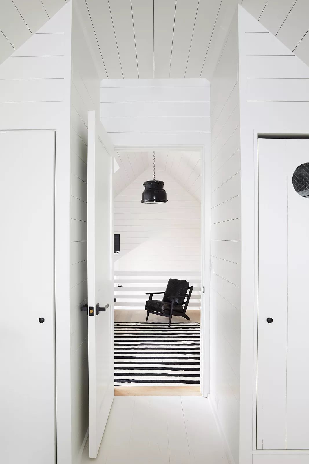 Hallway with white shiplap walls, black rocking chair on striped rug, and doors to storage.