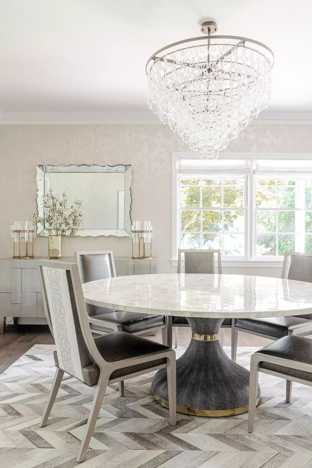 Dining room with round marble table, upholstered chairs, mirror, patterned rug, and chandelier.