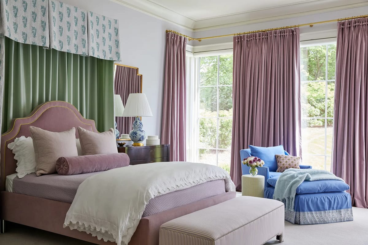 Bedroom with pink bed, blue armchair, large windows, curtains, and decorative pillows.