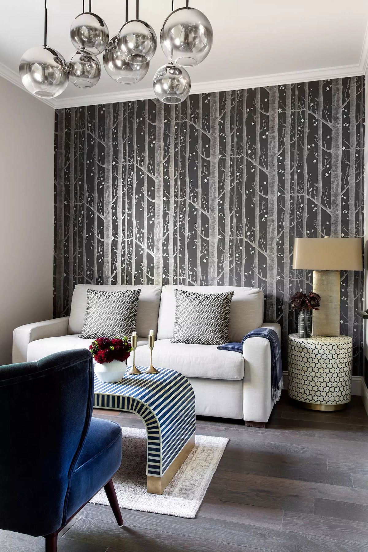 Living room with white sofa, blue armchair, striped ottoman, and tree-patterned wallpaper.