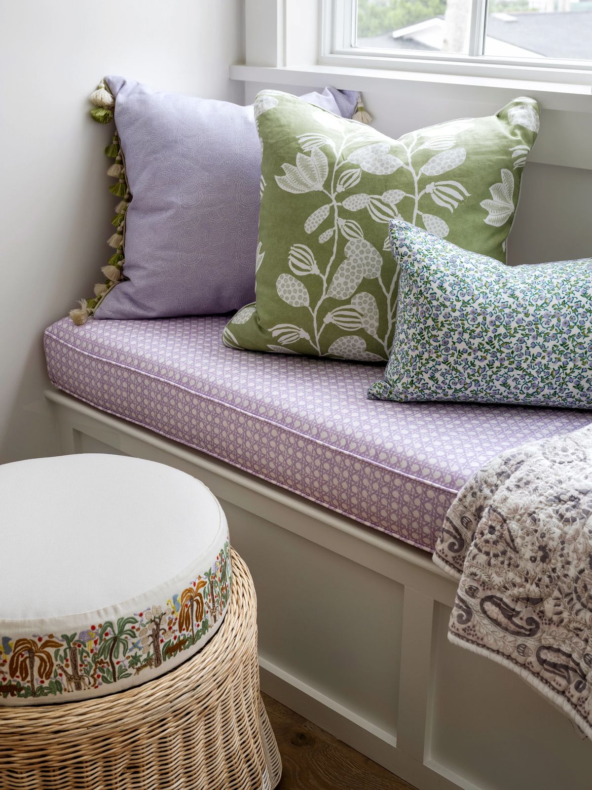 Window seat area with purple cushion, decorative pillows, and a round woven pouf.