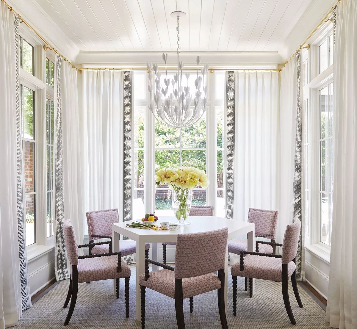 Dining room with white table, pink upholstered chairs, floral arrangement, windows, and light fixture