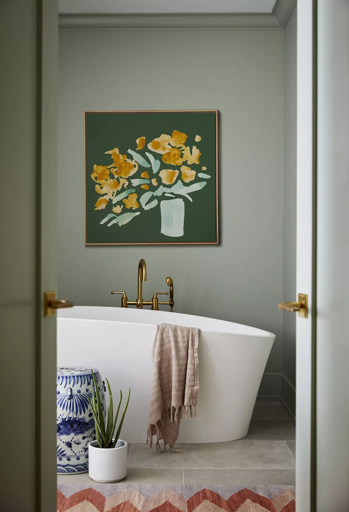 Bathroom with freestanding tub, gold faucet, floral painting, patterned rug, and potted plant.