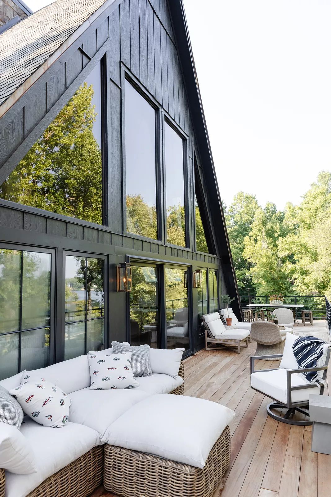 Deck with sectional sofa, wicker chair, metal chair, large windows, and wooden flooring surrounded by greenery