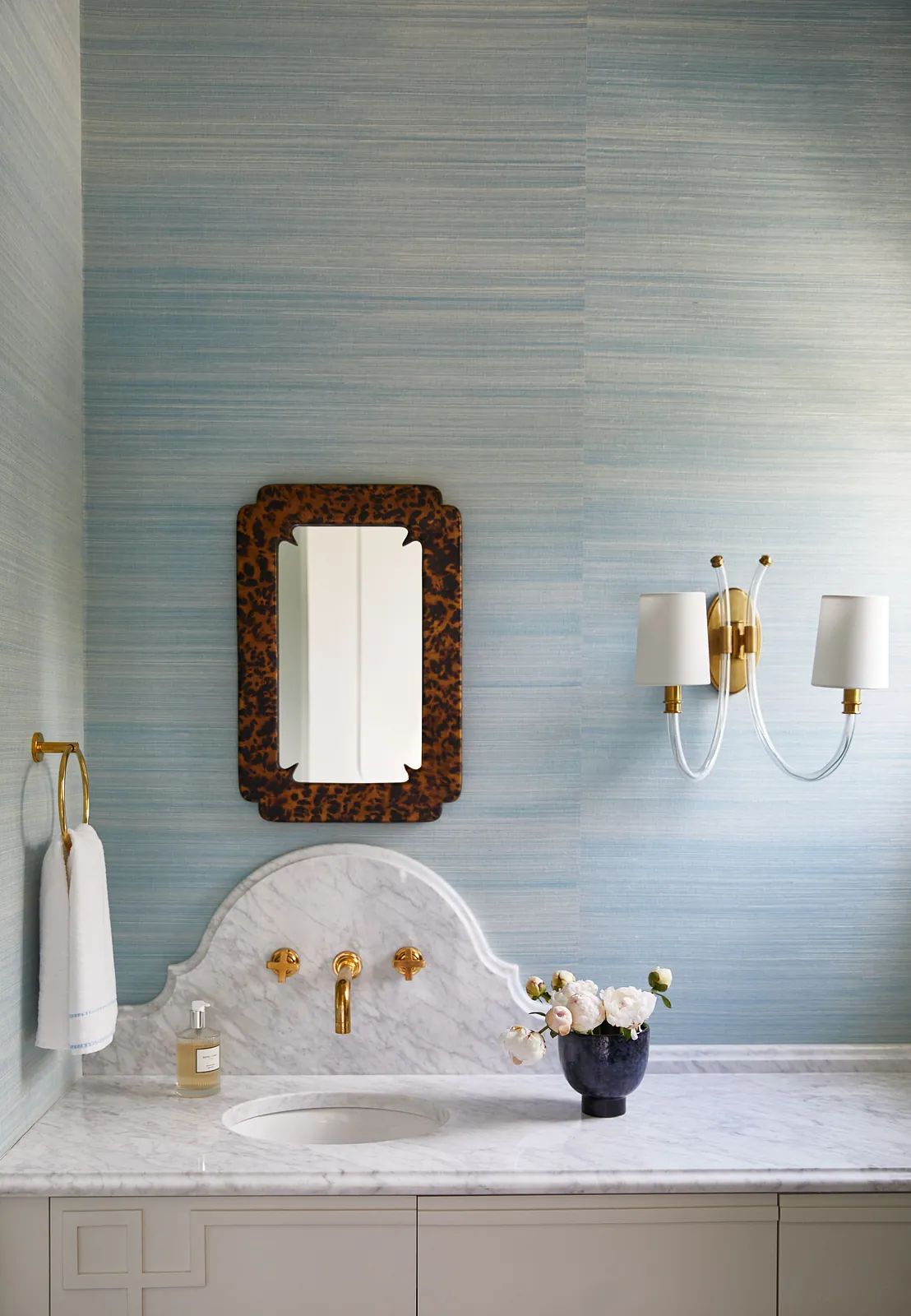 Bathroom with marble countertop, wall-mounted mirror, wall sconce, and floral decor on the sink.