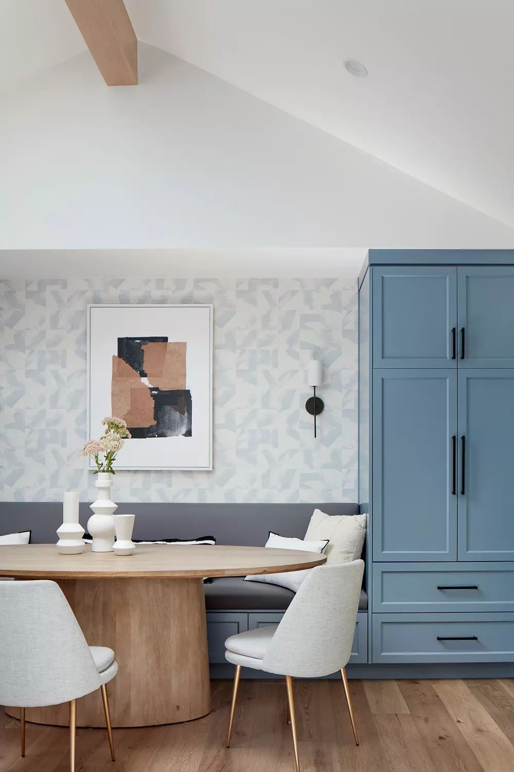Dining area with round wooden table, light gray chairs, blue built-in cabinet, geometric wallpaper, and abstract artwork