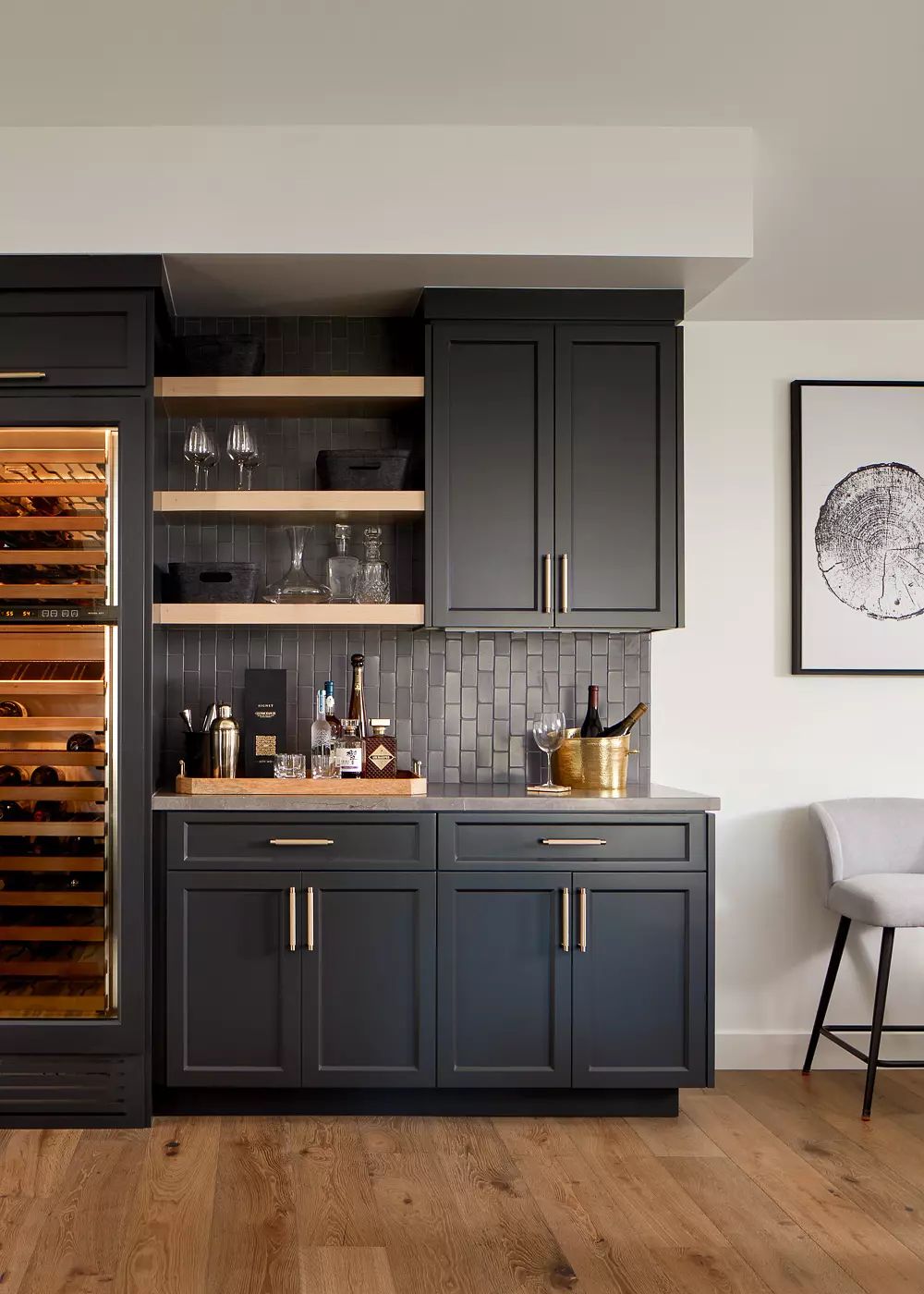 Kitchen with dark cabinetry, wine fridge, drink station, wooden shelving, and bar stool on light wood flooring.