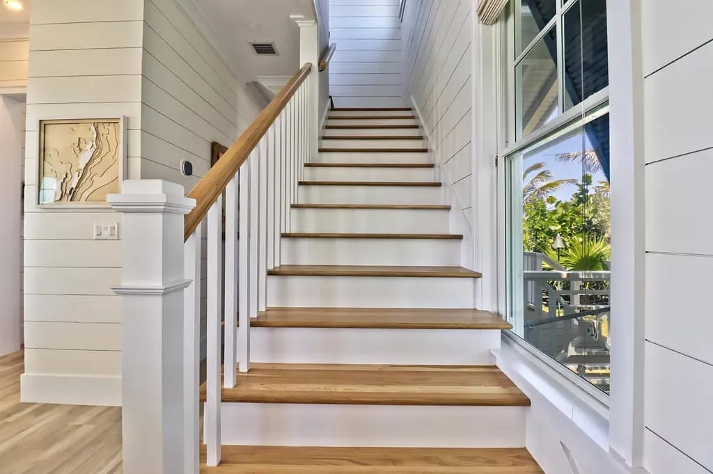 Hallway with wooden staircase, shiplap walls, large window, and wall art.